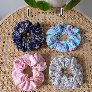 Op de afbeelding: Vier bloemen scrunchies in marineblauw, lichtblauw, roze en lavendel. Elke scrunchie heeft een ander bloemenpatroon. De scrunchies zijn gerangschikt op een geweven oppervlak, met de letters I, J, K en L ernaast.