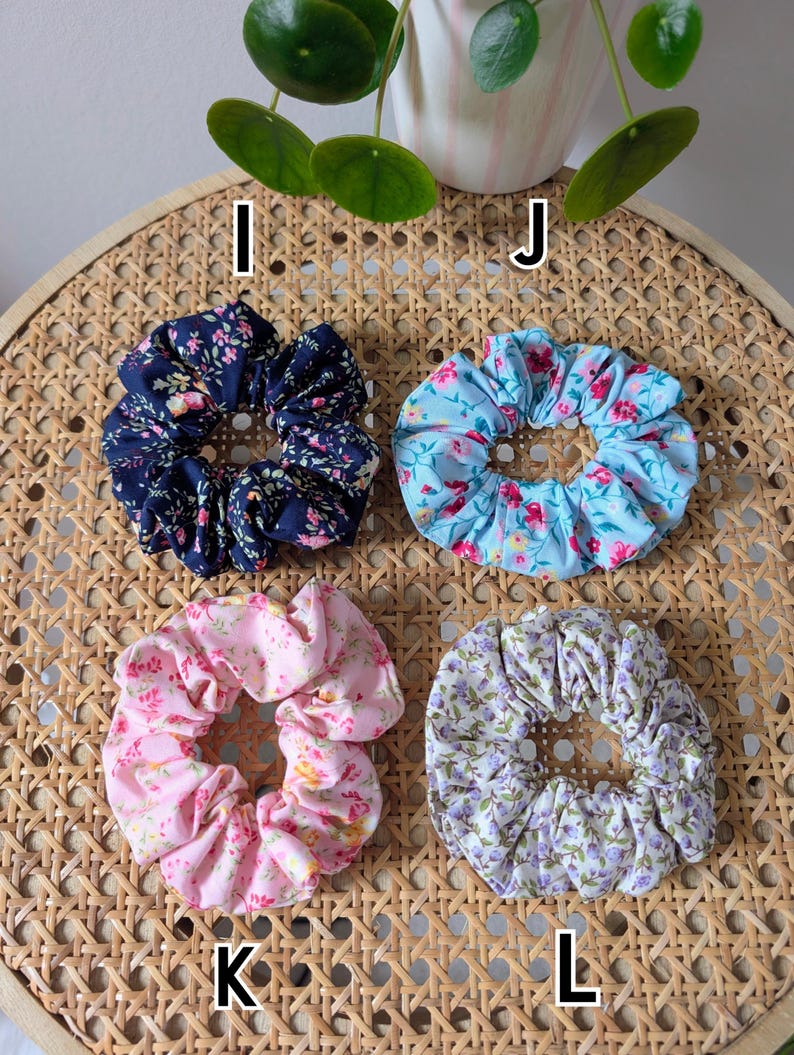 Op de afbeelding: Vier bloemen scrunchies in marineblauw, lichtblauw, roze en lavendel. Elke scrunchie heeft een ander bloemenpatroon. De scrunchies zijn gerangschikt op een geweven oppervlak, met de letters I, J, K en L ernaast.