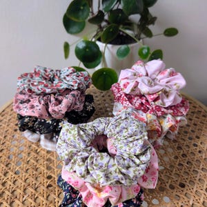 Op de afbeelding: Stapels bloemenstof scrunchies in verschillende kleuren, waaronder roze, rood, paars en marineblauw. De scrunchies zijn gerangschikt op een geweven oppervlak, met een potplant op de achtergrond. Haaraccessoires.