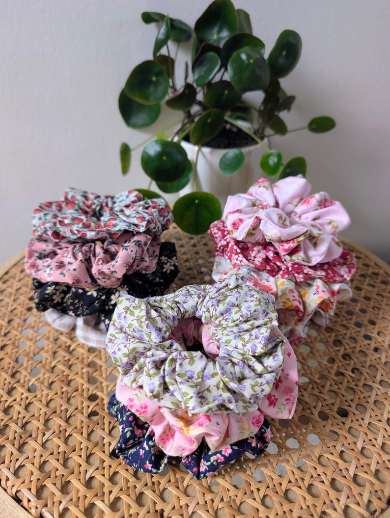 Op de afbeelding: Stapels bloemenstof scrunchies in verschillende kleuren, waaronder roze, rood, paars en marineblauw. De scrunchies zijn gerangschikt op een geweven oppervlak, met een potplant op de achtergrond. Haaraccessoires.