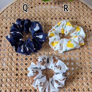 Op de afbeelding: Drie stoffen scrunchies met verschillende patronen. E&eacute;n is marineblauw met witte eenden en gele bloemen, een andere is wit met gele eenden en de derde is wit met bruine luiaards. Ze liggen op een geweven oppervlak.