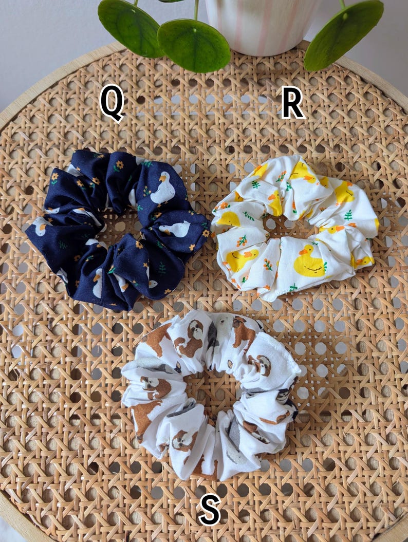 Op de afbeelding: Drie stoffen scrunchies met verschillende patronen. E&eacute;n is marineblauw met witte eenden en gele bloemen, een andere is wit met gele eenden en de derde is wit met bruine luiaards. Ze liggen op een geweven oppervlak.