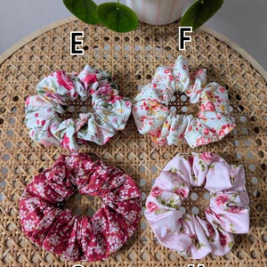 Op de afbeelding: Vier bloemen scrunchies in verschillende kleuren: wit met roze en rode bloemen, wit met oranje en rode bloemen, bordeauxrood met witte bloemen en roze met roze en groene bloemen. Op een geweven oppervlak.