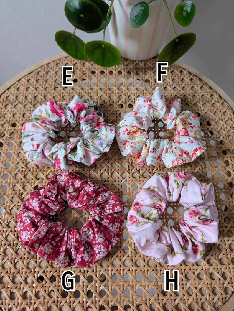 Op de afbeelding: Vier bloemen scrunchies in verschillende kleuren: wit met roze en rode bloemen, wit met oranje en rode bloemen, bordeauxrood met witte bloemen en roze met roze en groene bloemen. Op een geweven oppervlak.