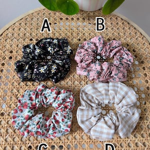 Op de afbeelding: Vier stoffen scrunchies met verschillende patronen worden getoond op een geweven oppervlak. De scrunchies bevatten een zwarte bloemenprint, een roze bloemenprint, een aardbeienprint en een bruin en wit geruit patroon. De scrunchies zijn gelabeld A, B, C en D.