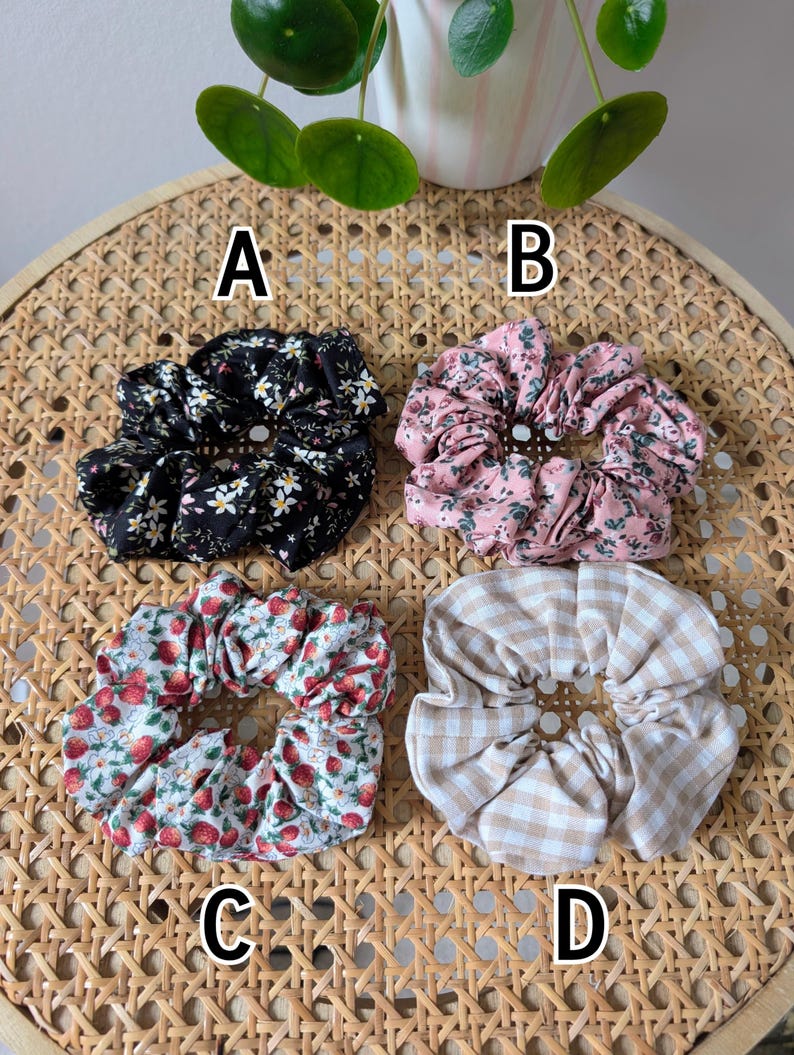 Op de afbeelding: Vier stoffen scrunchies met verschillende patronen worden getoond op een geweven oppervlak. De scrunchies bevatten een zwarte bloemenprint, een roze bloemenprint, een aardbeienprint en een bruin en wit geruit patroon. De scrunchies zijn gelabeld A, B, C en D.