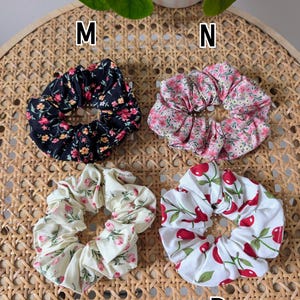 Op de afbeelding: Vier stoffen scrunchies met bloemen- en fruitpatronen. E&eacute;n is zwart met roze en gele bloemen, een andere is roze met witte bloemen, een derde is cr&egrave;me met roze bloemen en de laatste is wit met rode kersen. De scrunchies liggen op een geweven oppervlak.