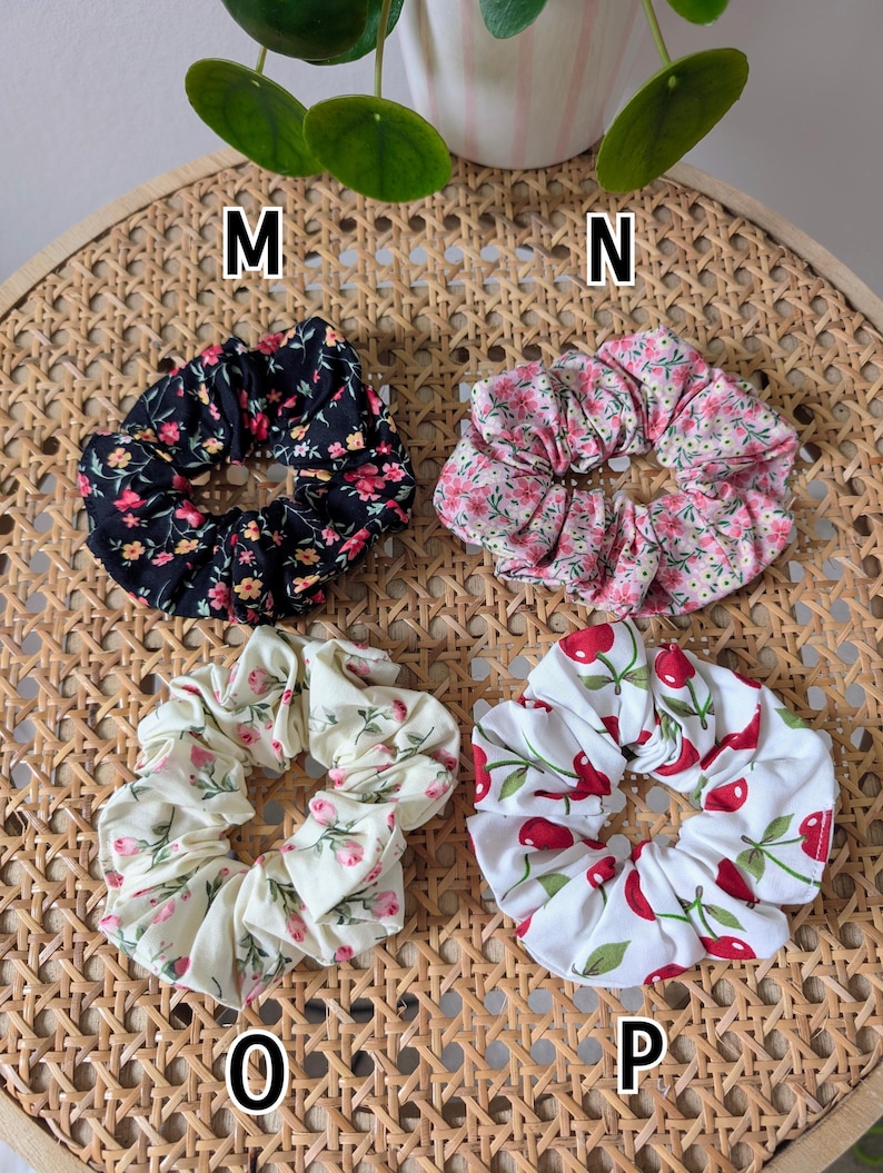 Op de afbeelding: Vier stoffen scrunchies met bloemen- en fruitpatronen. E&eacute;n is zwart met roze en gele bloemen, een andere is roze met witte bloemen, een derde is cr&egrave;me met roze bloemen en de laatste is wit met rode kersen. De scrunchies liggen op een geweven oppervlak.