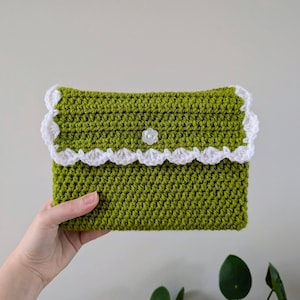 May include: A handmade, olive green crochet clutch with a white scalloped trim and a small white flower button closure. The rectangular bag is held up against a neutral background.