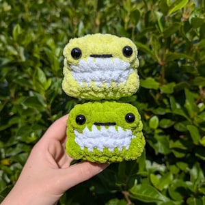 May include: Two handmade crochet frog plushies. The top frog is light yellow, and the bottom frog is green. Both have black eyes and white mouths. The plushies are held in front of a background of green leaves.