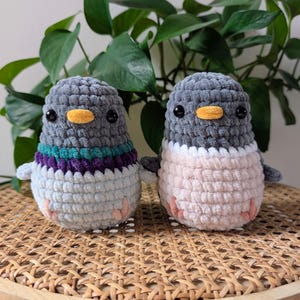 May include: Two handmade crochet pigeon plushies. One has a gray head, a purple and teal stripe, and a light blue body. The other has a gray head and a pink body. Both have black eyes and orange beaks. They are sitting on a woven surface.