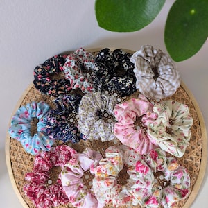Op de afbeelding: Een verzameling scrunchies met bloemenpatronen in verschillende kleuren, waaronder zwart, roze, blauw en wit. De scrunchies zijn gerangschikt op een geweven dienblad, met een verscheidenheid aan stoffen ontwerpen en texturen. Haaraccessoire.