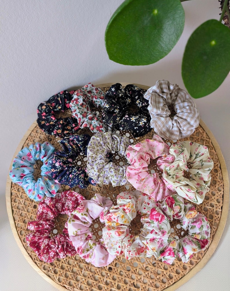 Op de afbeelding: Een verzameling scrunchies met bloemenpatronen in verschillende kleuren, waaronder zwart, roze, blauw en wit. De scrunchies zijn gerangschikt op een geweven dienblad, met een verscheidenheid aan stoffen ontwerpen en texturen. Haaraccessoire.