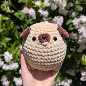 May include: A round, beige crochet plush toy shaped like a pug. It features dark brown floppy ears, a dark brown snout, two black plastic eyes, and a small pink tongue sticking out. The texture is soft and fuzzy, created with thick yarn.