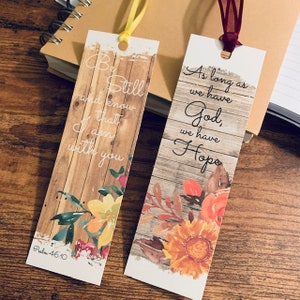 Può includere: Due segnalibri floreali con citazioni ispiratrici. Il primo segnalibro presenta uno sfondo in legno con la citazione "Stai calmo e sappi che io sono con te" e il riferimento biblico "Salmo 46:10". Il secondo segnalibro presenta uno sfondo in legno con la citazione "Finché abbiamo Dio, abbiamo speranza".
