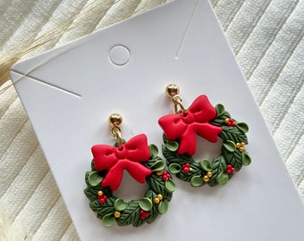 May include: A pair of green and red Christmas wreath earrings with a red bow. The earrings are made of polymer clay and have gold hooks.