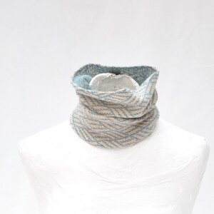 May include: A knitted cowl scarf in a neutral beige and light blue chevron pattern. The scarf is displayed on a white mannequin bust. The interior of the cowl is a solid light blue color. This neck warmer is a cozy winter accessory.