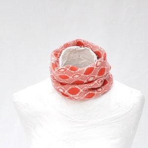 Puede incluir: Bufanda infinita con estampado rojo y blanco, colocada sobre un busto de maniquí blanco. La bufanda presenta un diseño ovalado repetido. La tela parece tejida, sugiriendo un accesorio cálido. Circunferencia aproximada de 150 cm.