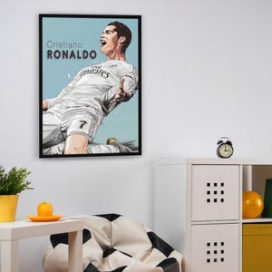 May include: A framed poster featuring a digital illustration of Cristiano Ronaldo in a white football shirt with the text "Cristiano Ronaldo" and "Fly Emirates" on the shirt. The background is a light blue colour.