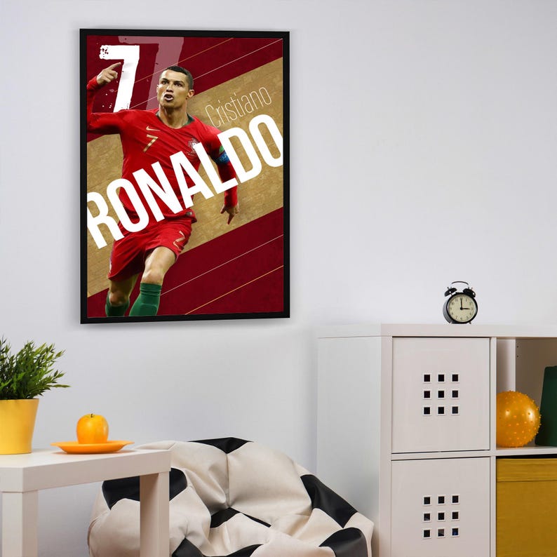 Pu&ograve; includere: Un poster incorniciato con una foto di Cristiano Ronaldo in una divisa da calcio rossa e verde, che celebra un gol. Il poster ha uno sfondo rosso e oro con strisce diagonali bianche e il testo "Cristiano Ronaldo" in lettere bianche.