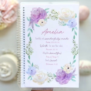 Puede incluir: Un cuaderno de espiral con un borde floral de acuarela en tonos morado, crema y verde. La portada muestra el nombre Amelia y varios versículos bíblicos, incluyendo Fearfully and wonderfully made y Worth far more than rubies.