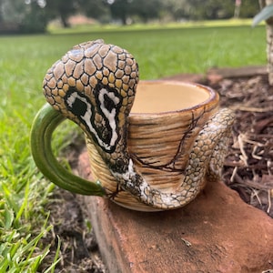 Könnte beinhalten: Ein Keramikbecher mit einem geformten Kobra-Henkel und Körper. Die Schlange hat ein texturiertes, hellbraunes und braunes Schuppenmuster mit einem grünen Griffbereich. Der Becher hat ein hellbeiges Inneres und ein braunes, holzartiges Äußeres.