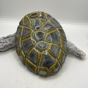 May include: A ceramic turtle shell with a textured surface. The shell is primarily blue-grey with accents of gold and brown. The shell is resting on a piece of driftwood.