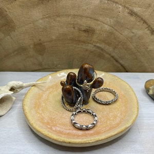 May include: A ceramic ring holder shaped like a cactus with a brown and orange glaze. The holder has three prongs for rings and is sitting on a light orange dish.