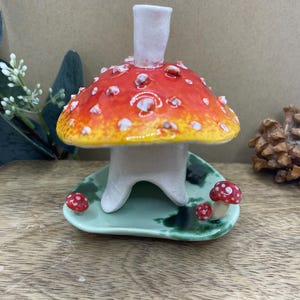 Op de afbeelding: Een grillige keramische paddenstoel kaarsenhouder. De paddenstoel heeft een rode hoed met witte stippen en een gele onderkant. Hij staat op een lichtgroene, bladvormige basis, met kleine rode paddenstoelen. De kaarsenhouder is wit.