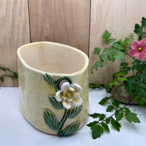 Puede incluir: Una maceta de cerámica con un esmalte amarillo y una flor blanca esculpida en el lateral. La maceta tiene forma ovalada y una superficie texturizada.