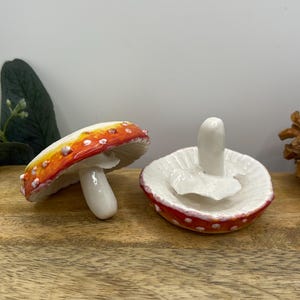 May include: Two ceramic mushroom-shaped trinket dishes. One is a cap with a stem, the other is a cap with a small stem. The caps are red with white spots and a yellow-orange gradient. The stems are white.
