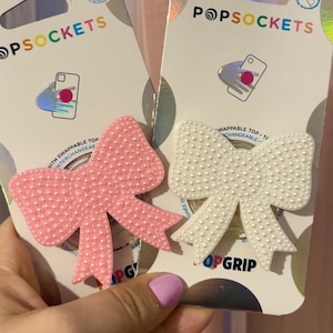 Pode incluir: Dois suportes para telefone PopSockets, um rosa e outro branco, com textura perolada e design de laço. O texto "PopSockets" e "PopGrip" é visível na embalagem.