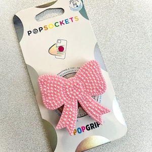 May include: Pink PopGrip phone grip with a pearl-like texture in the shape of a bow. The packaging features the PopSockets logo and the text "Swappable Top -  The Top is Changeable".