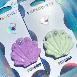 Può includere: Due PopSockets PopGrip per telefono a forma di conchiglia con dettagli di perle. Uno è viola chiaro e l'altro è verde chiaro.