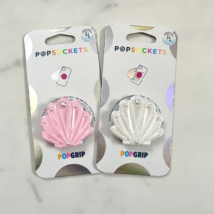 Peut inclure: Deux poignées de téléphone en forme de coquille. La poignée de gauche est rose avec des accents de perles blanches. La poignée de droite est blanche avec des accents de perles blanches. Les deux poignées ont les mots "PopGrip" imprimés dessus.