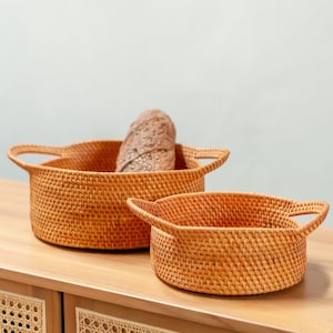 Op de afbeelding: Twee geweven rotan manden met handvatten. Eén mand bevat een brood. De manden hebben een warme, natuurlijke kleur en staan op een houten oppervlak. De manden zijn leeg, behalve het brood.