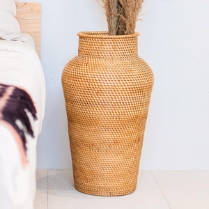 Puede incluir: Un jarrón grande de mimbre tejido natural con una boca ancha y un cuerpo redondeado. El jarrón está hecho de un material marrón claro y tiene un aspecto natural y rústico.