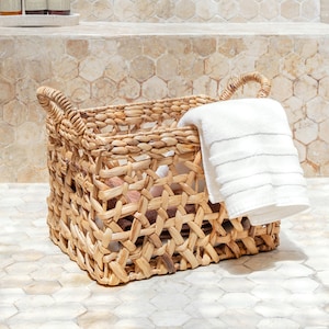 May include: A woven wicker basket with a white towel inside. The basket is sitting on a tiled floor in a bathroom setting.