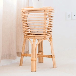 Peut inclure: Un support de plante en rotin naturel avec un design cylindrique. Le support présente un dessus en forme de panier tressé et une base à trois pieds. Un pot en céramique blanche avec des plantes séchées est posé sur le support. Le support mesure environ 76 cm de haut.