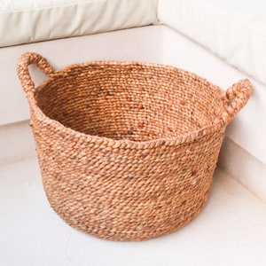 Puede incluir: Una cesta grande, redonda y tejida con dos asas. La cesta está hecha de materiales naturales y tiene un color marrón claro.