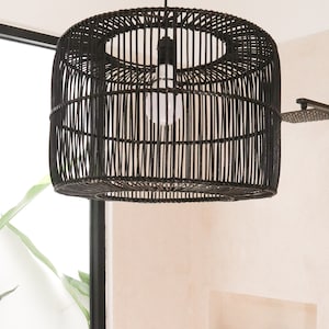Puede incluir: Lámpara colgante de mimbre negro tejido colgada del techo de un baño. La lámpara es redonda y tiene una bombilla blanca en su interior.