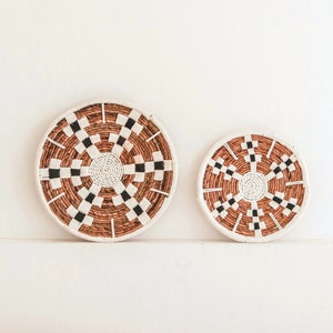 May include: Two woven wall decor pieces with a black and white checkered pattern on a brown background. The larger piece is on the left and the smaller piece is on the right. Both pieces are hanging on a white wall above a brown couch.