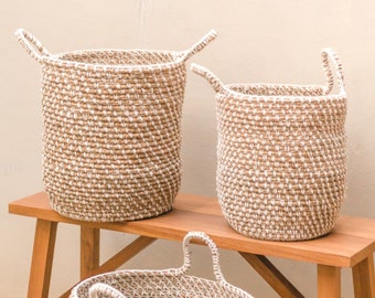 May include: Three woven baskets with white accents and handles sit on a wooden bench. The baskets vary in size, with the largest on the left and the smallest in the center. The baskets are made of a natural fiber.