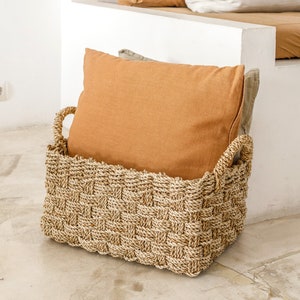 Könnte beinhalten: Ein geflochtener Seegras-Korb mit zwei braunen Kissen darin. Der Korb steht auf einem hellbeigen Fliesenboden.