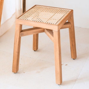 Puede incluir: Un taburete cuadrado de madera con un asiento de ratán tejido. El taburete tiene un acabado de madera natural y cuatro patas.