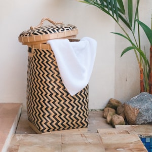 May include: A black and brown woven bamboo laundry basket with a white towel inside. The basket is sitting on a wooden surface next to a potted palm plant.