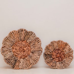 May include: Two woven wall decor pieces in a flower shape. The larger piece is approximately 20 inches in diameter, and the smaller is about 14 inches. Both are made of natural-colored woven material with a darker center.