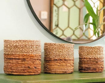 May include: Three woven baskets with a natural brown and tan color. The baskets are made of natural materials and have a rustic look. They are sitting on a green surface.