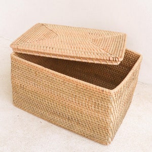 以下が含まれることがあります： 薄い茶色の籐でできた、蓋付きの自然な織り収納バスケット。蓋が開いており、バスケットは空です。