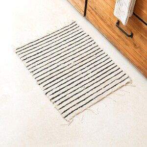 May include: A white and black striped rug with frayed edges. The rug is made of a natural fiber and has a simple, minimalist design.